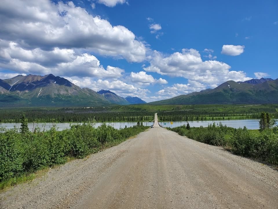 Gravel road crossing a bridge on the Denali Highway in Alaska, with green hills and distant mountains beneath a cloud-filled sky